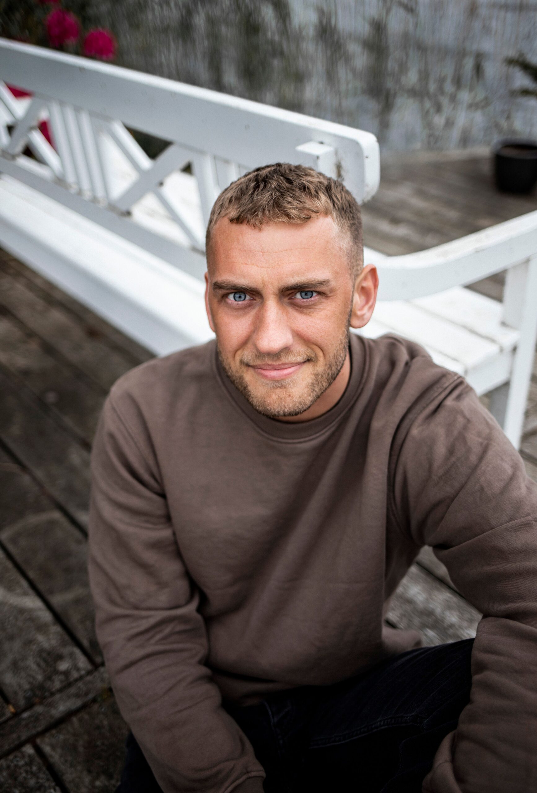 A relaxed man with short hair and blue eyes smiling while sitting outdoors on a white bench.