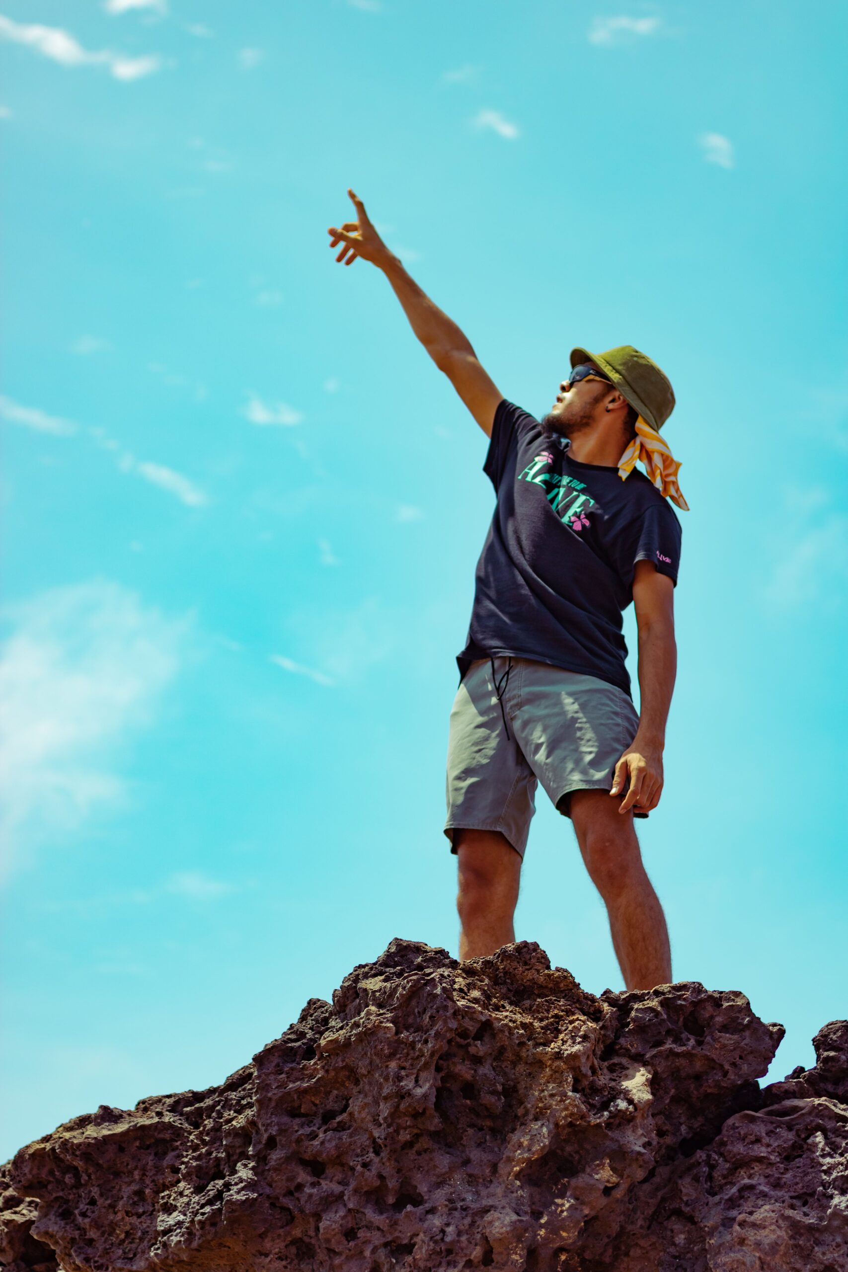 A man on a rocky peak wearing a hat points towards the sky under a bright blue sky.