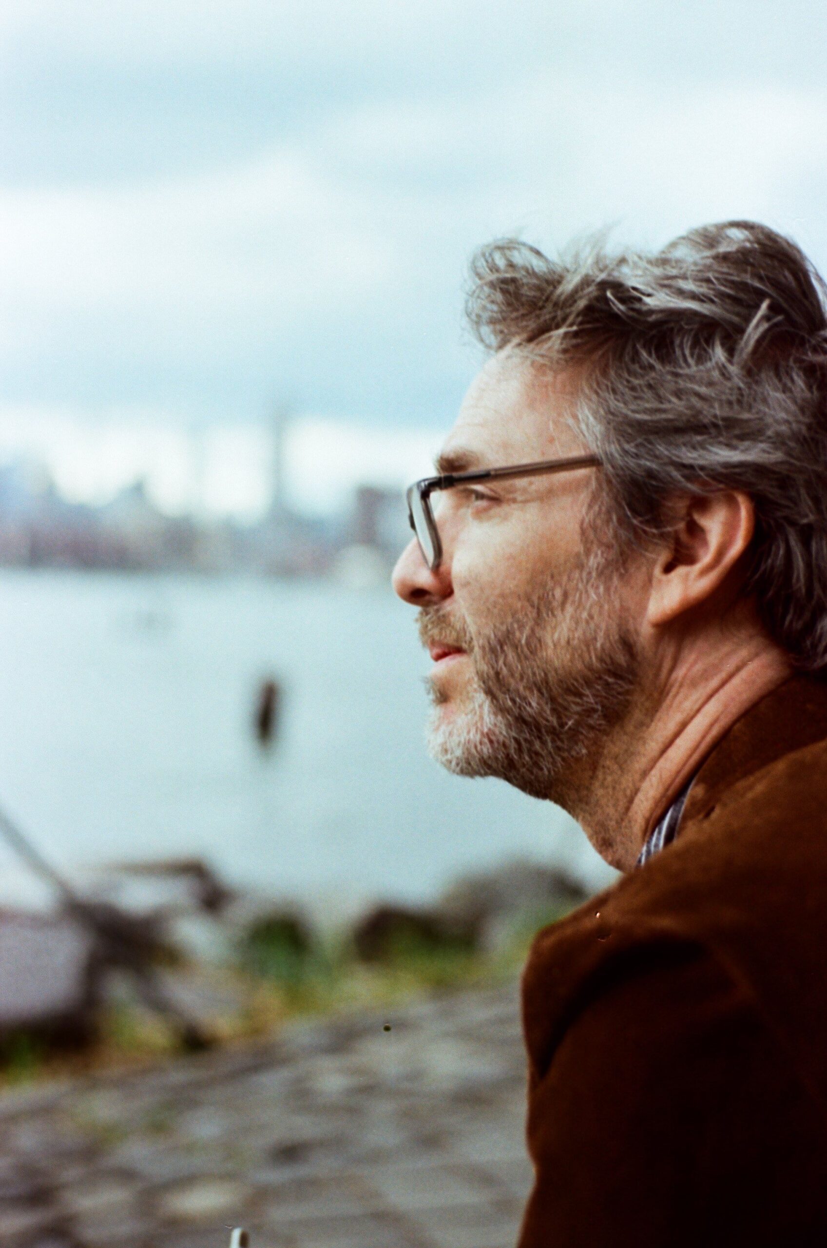 Bearded man in glasses gazing thoughtfully across New York City waterfront.