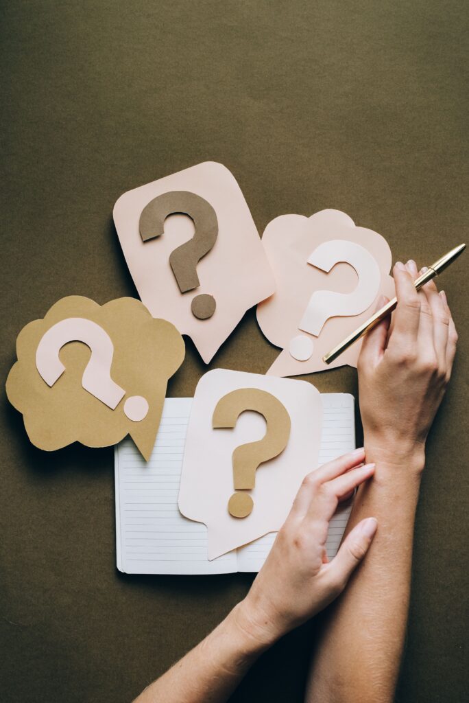 Hands holding pen next to paper question marks on notebook, symbolizing inquiry and creativity.