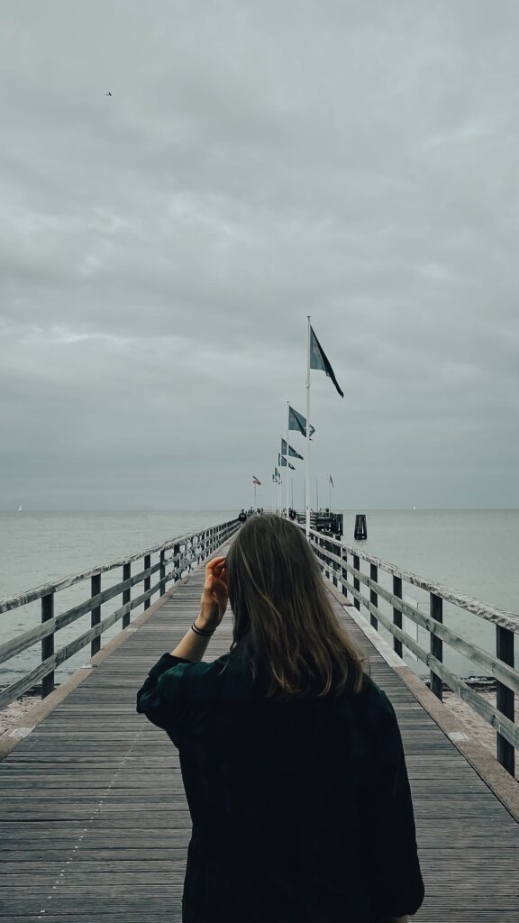 A person walks on a wooden pier extending into a calm sea under a cloudy sky.